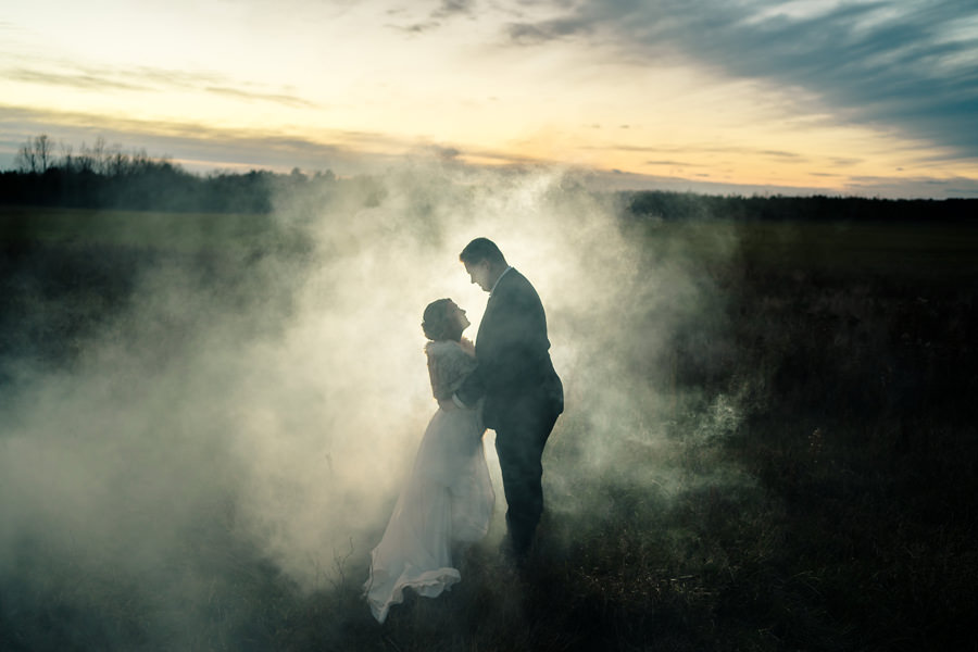 A bride and groom standing in a field with smoke in the background.