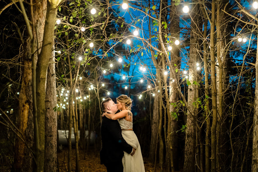 A bride and groom kiss under string lights in the woods.