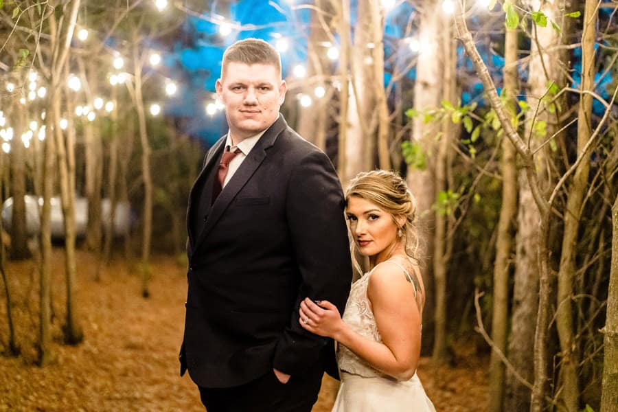 A bride and groom standing in a wooded area with string lights.