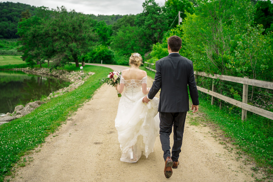 A bride and groom walking down a dirt path near a pond.