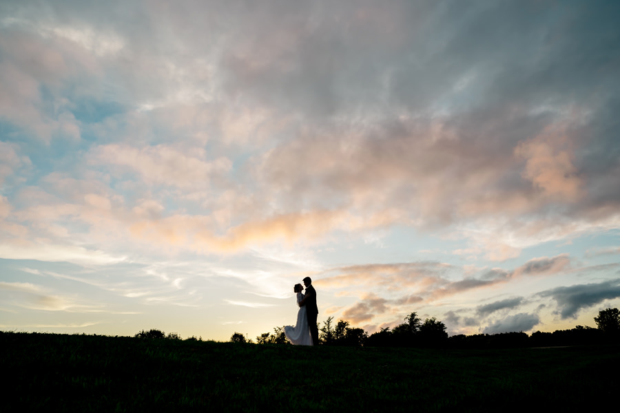 A bride and groom standing on a grassy hill at sunset.