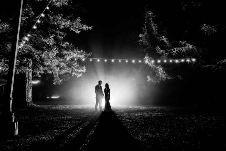A black and white photo of a bride and groom standing under string lights.