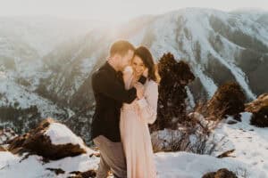 A couple embraces on top of a snowy mountain during their engagement session, captured for the Ultimate Wedding Planning Guide.