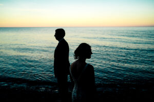 A bride and groom standing on the beach at sunset.