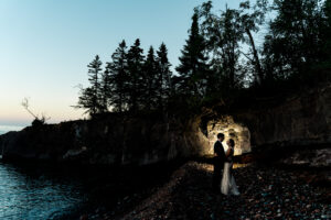 A bride and groom standing in front of a cave at dusk.