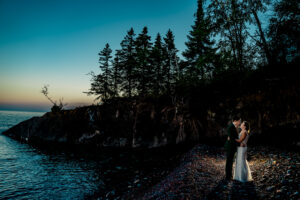 A bride and groom standing on a rocky shore at sunset.