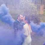Courtney and James' Love-Filled Backyard Trempleau Wedding showcases a bride and groom kissing amidst a blue smoke cloud.