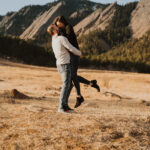 A couple is hugging in the middle of a field with mountains in the background.