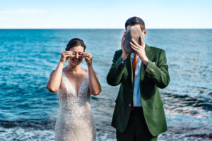A bride and groom standing on the beach with their hands in their pockets.