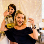 Two women displaying celebration at Courtney and James’ Love-Filled Backyard Trempleau Wedding, with a bottle of liquor on a table.