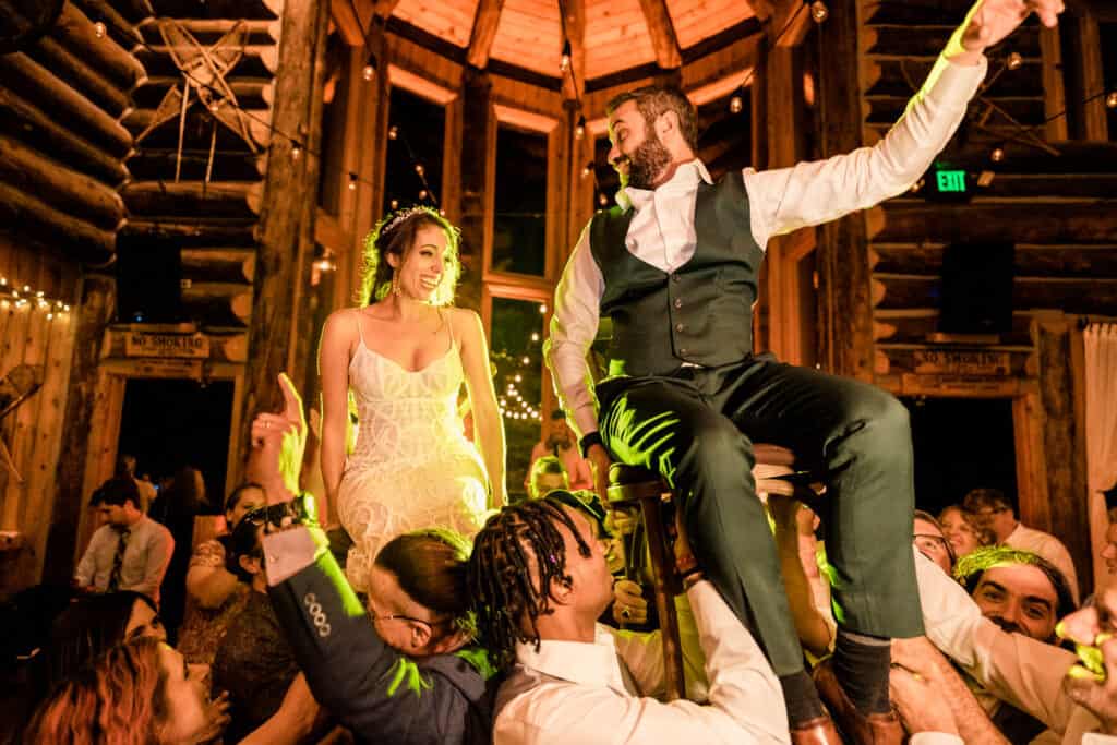 A man and woman sitting on a stool in a room with people.