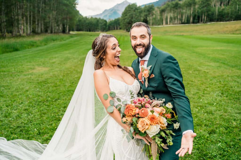 A bride and groom laughing in a field with mountains in the background.
