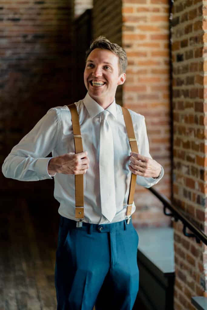 A man wearing suspenders and a tie in front of a brick wall.