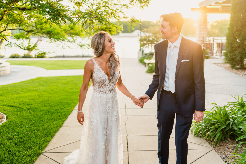 A bride and groom holding hands on a sidewalk in front of a lake.