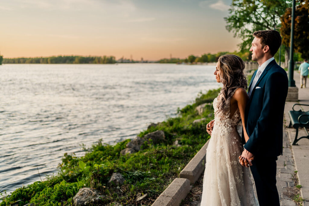 A bride and groom standing by a river at sunset.