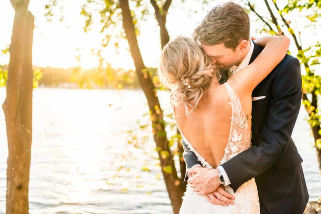 A bride and groom embrace in front of a lake at sunset.