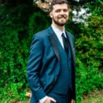 A groom in a tuxedo smiling in front of bushes.