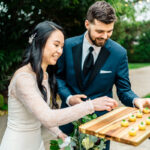 A bride and groom serving appetizers on a wooden board.