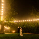 A bride and groom standing under string lights at night.