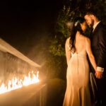 A bride and groom standing in front of a fire pit.