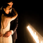 A bride and groom embracing in front of a fire.