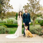 A bride and groom pose with their dog in a garden.