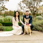 A bride and groom pose with their dog in a garden.