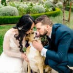 A bride and groom kissing their dog in front of a garden.