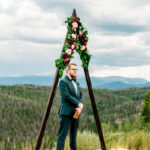 A groom standing in front of a wedding arch in the mountains.