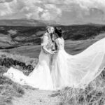 Two brides kissing on a hillside in a black and white photo.