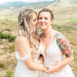 Two brides hugging in a field with mountains in the background.