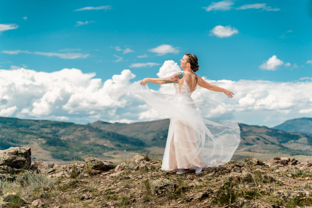 A bride is standing on top of a mountain with her veil in the air.