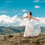 A bride is standing on top of a mountain with her veil in the air.