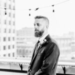 A man in a suit standing on a balcony with a beard.