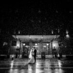 A bride and groom standing in front of a building in the rain.