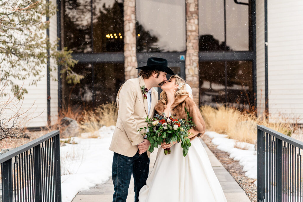 A bride and groom kiss in front of a building in the snow.