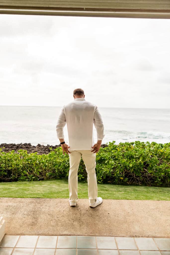 A man in a white outfit stands on a patio facing the ocean, with greenery in the foreground and a cloudy sky in the background.