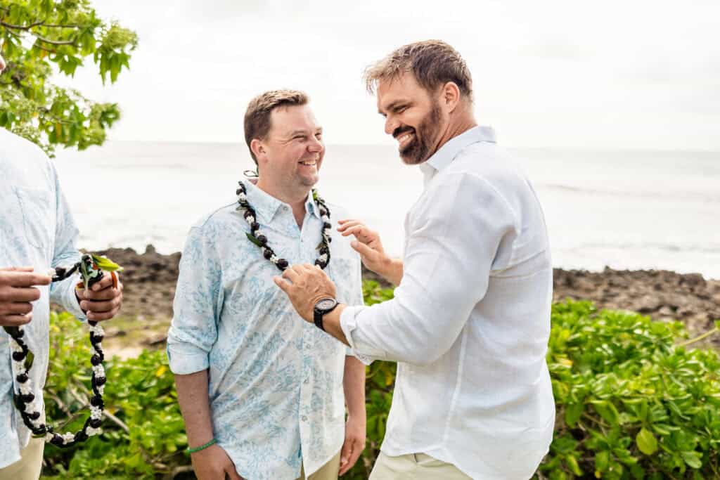 Two men in white shirts smiling and standing by the ocean, one placing a lei around the other's neck. Verdant plants and the sea are in the background.