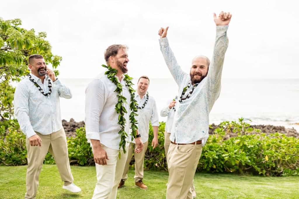 Four men wearing white shirts and beige pants, with two of them adorned with leafy garlands, stand together on grassy terrain by the sea. One man has his arms raised in celebration.