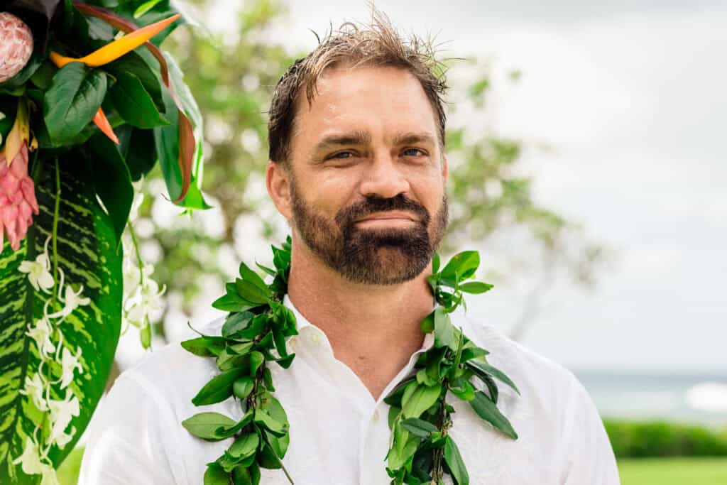 A man with a beard wears a white shirt and a green lei, standing outdoors with foliage and a blurred background.