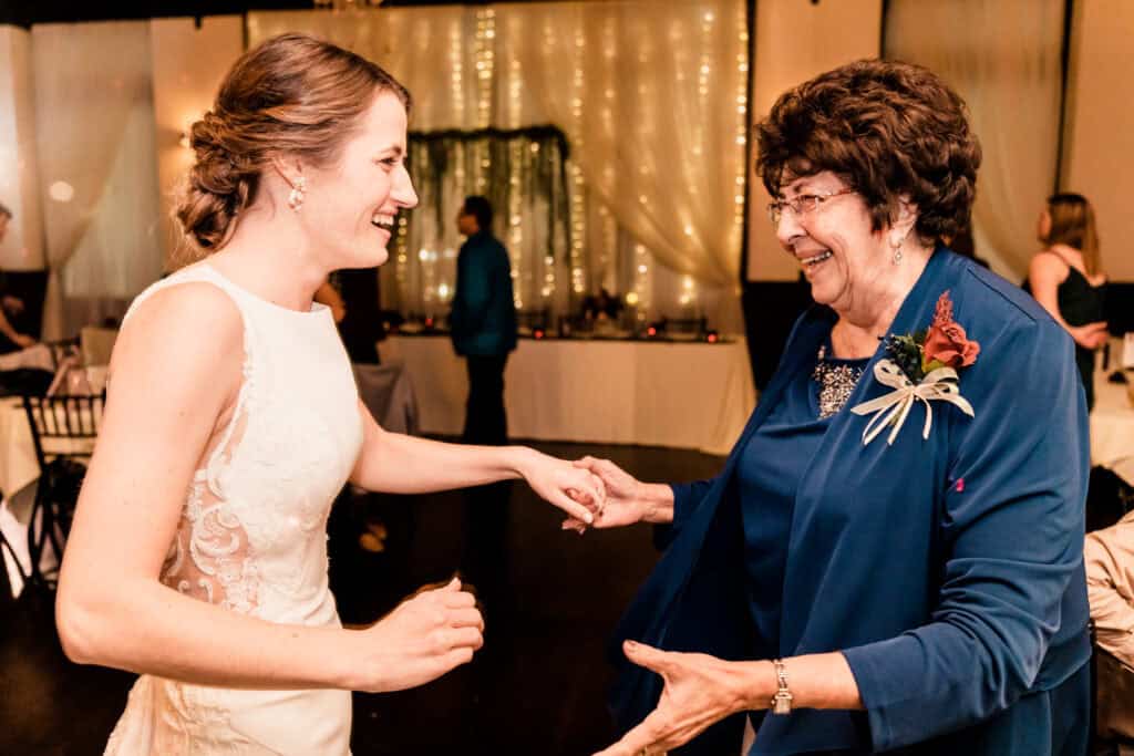 A bride in a white dress and an older woman in a blue dress share a joyful moment dancing together at a wedding reception.
