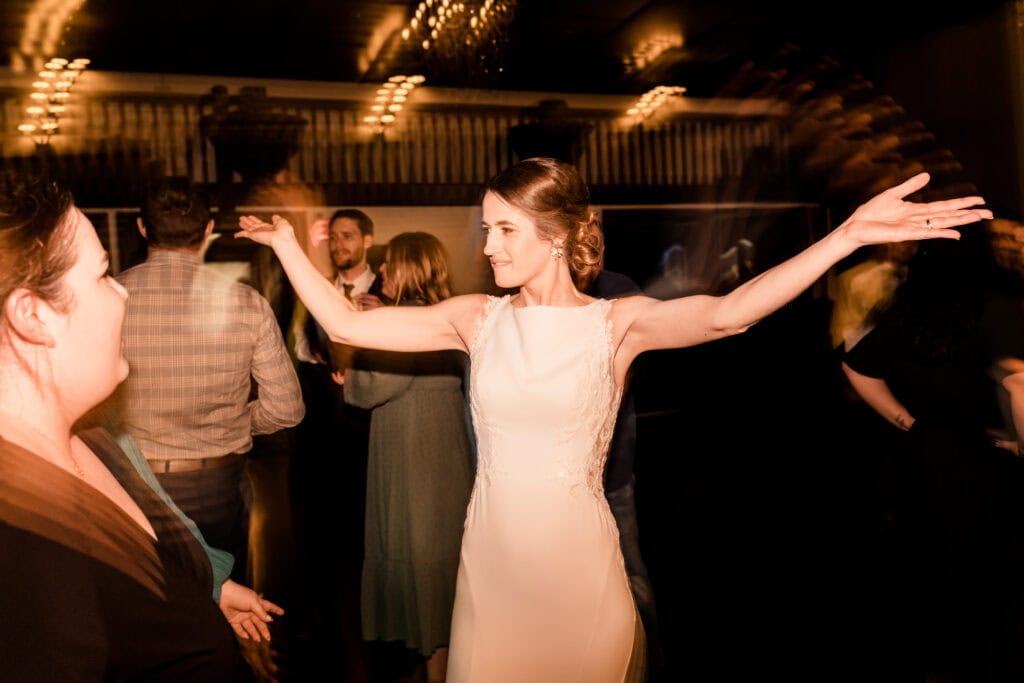 A woman wearing a white dress stands with her arms outstretched, smiling, in a dimly lit room with other people around.