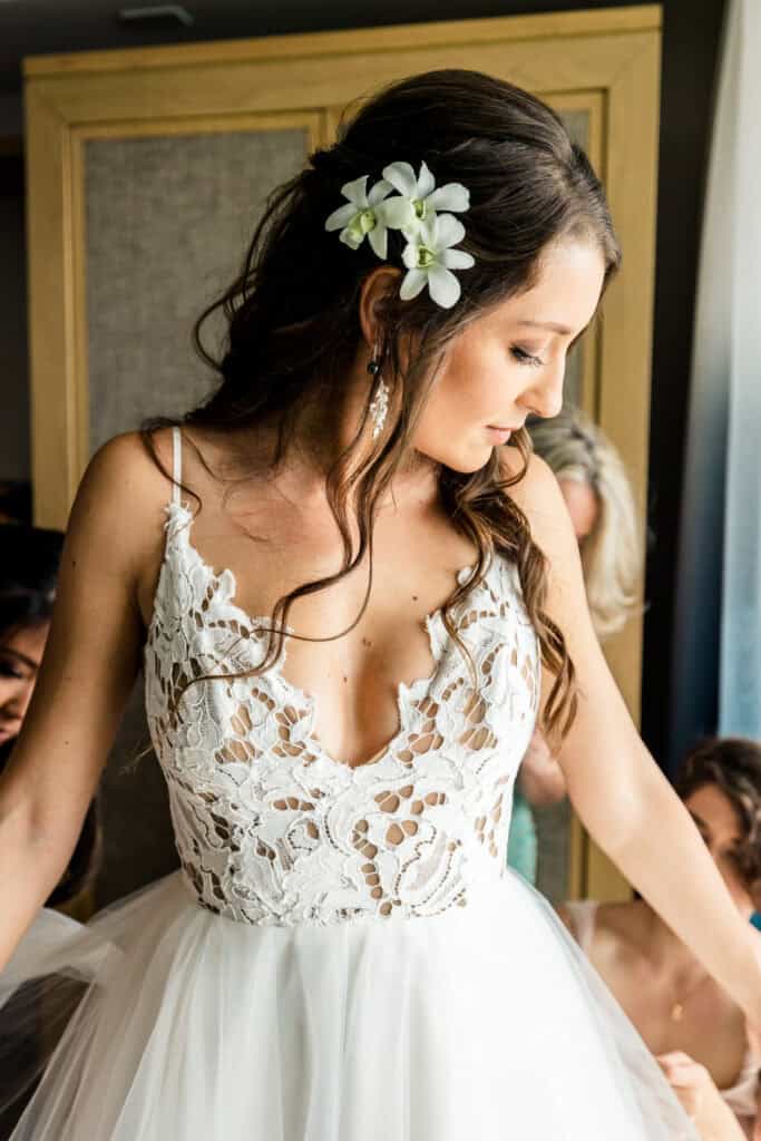 A bride with long hair adorned with white flowers, wearing a lace and tulle wedding dress, looks down as she prepares for her wedding day.