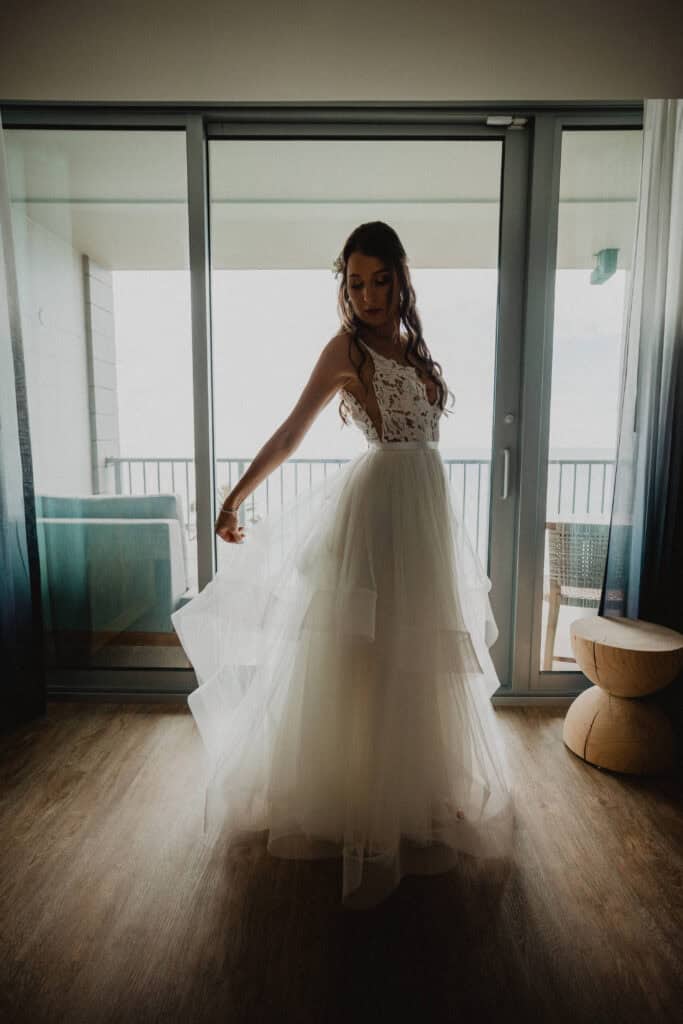 A woman in a white lace and tulle wedding dress stands inside near a balcony door, holding her dress and looking down.