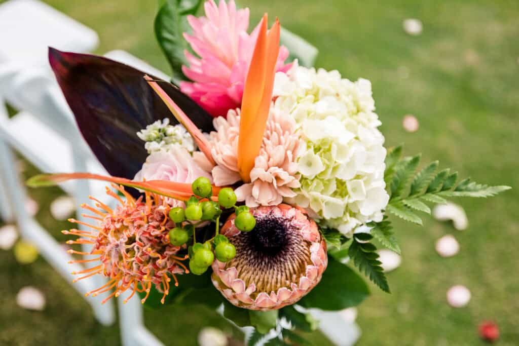 A close-up of a vibrant bouquet featuring a variety of flowers, including hydrangea, protea, and bird of paradise, set against a grassy background.