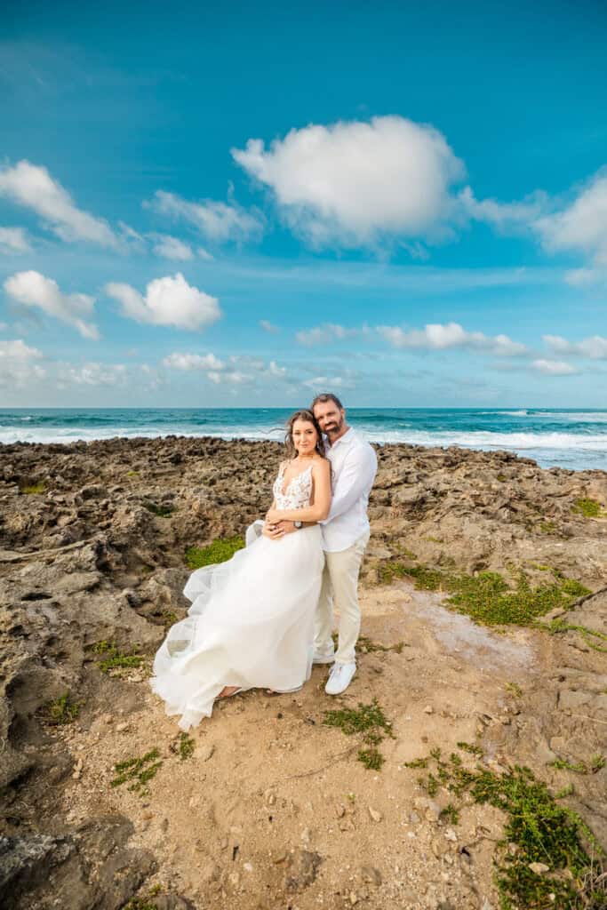 A couple dressed in wedding attire stands together on a rocky shoreline with the ocean and a blue sky with clouds in the background.