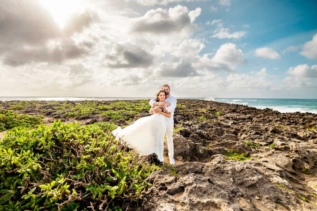 A couple in wedding attire embraces on a rocky coastal landscape under a partly cloudy sky, with greenery and ocean waves in the background.