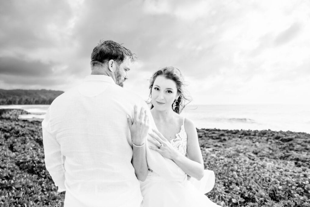 A bride in a white dress and a groom in a white shirt stand on a rocky shoreline with the ocean in the background under a cloudy sky. The bride looks at the camera while the groom looks away.