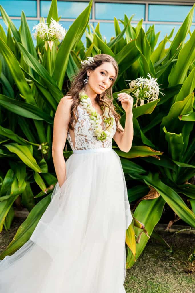 A woman in a white, lace bodice gown stands in front of large green plants with white flowers, wearing a floral lei and holding a flower to her hair.