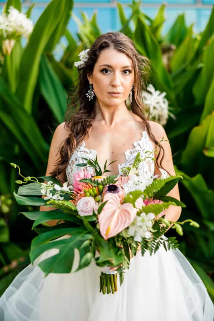 A bride in a white wedding dress holds a bouquet of various flowers and greenery, standing in front of large green foliage.